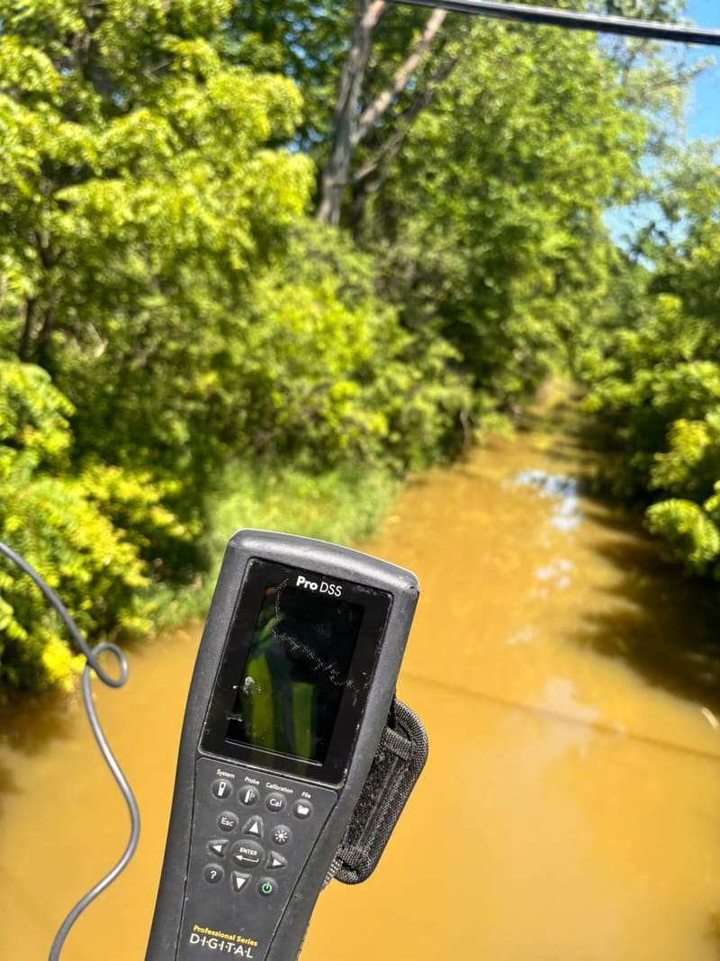 A handheld water quality meter held over a muddy, tree-lined tributary